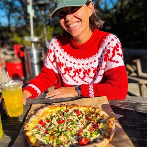 Puede incluir: Un su&eacute;ter rojo con un patr&oacute;n blanco, una gorra verde con texto naranja y una pizza con ingredientes est&aacute;n sobre una mesa de madera. Una persona sonr&iacute;e y sostiene una bebida con una pajita. La pizza tiene ingredientes rojos y verdes.