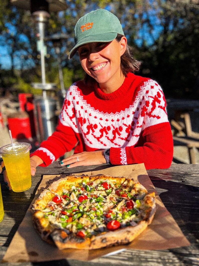 Puede incluir: Un su&eacute;ter rojo con un patr&oacute;n blanco, una gorra verde con texto naranja y una pizza con ingredientes est&aacute;n sobre una mesa de madera. Una persona sonr&iacute;e y sostiene una bebida con una pajita. La pizza tiene ingredientes rojos y verdes.
