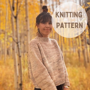May include: A woman wearing a light brown knitted jumper with a subtle speckled pattern. The jumper has a round neckline and long sleeves. The jumper is cropped and the woman is wearing it with a black skirt. The image is taken outdoors in a forest setting with autumn foliage in the background. The text "Knitting Pattern" is superimposed on the image.