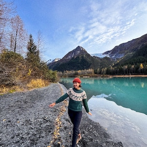 Puede incluir: Una persona con un su&eacute;ter verde y blanco estampado, un gorro rojo y pantalones oscuros camina por un sendero de grava junto a un lago turquesa. Monta&ntilde;as y &aacute;rboles se ven al fondo bajo un cielo azul.