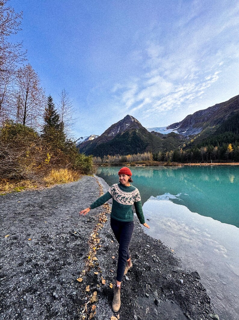 Puede incluir: Una persona con un su&eacute;ter verde y blanco estampado, un gorro rojo y pantalones oscuros camina por un sendero de grava junto a un lago turquesa. Monta&ntilde;as y &aacute;rboles se ven al fondo bajo un cielo azul.