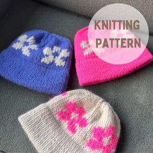 May include: Three knitted beanies in blue, pink, and cream, each with a floral design. The beanies are shown on a grey surface. The image includes the text "KNITTING PATTERN" within a white circle.