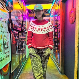 Puede incluir: Una persona con un su&eacute;ter rojo y blanco con tem&aacute;tica de esqu&iacute;, pantalones verdes con estampado floral y una gorra verde. La persona est&aacute; en un pasillo con luces de ne&oacute;n coloridas y obras de arte enmarcadas. Un letrero arriba dice "Ladies".