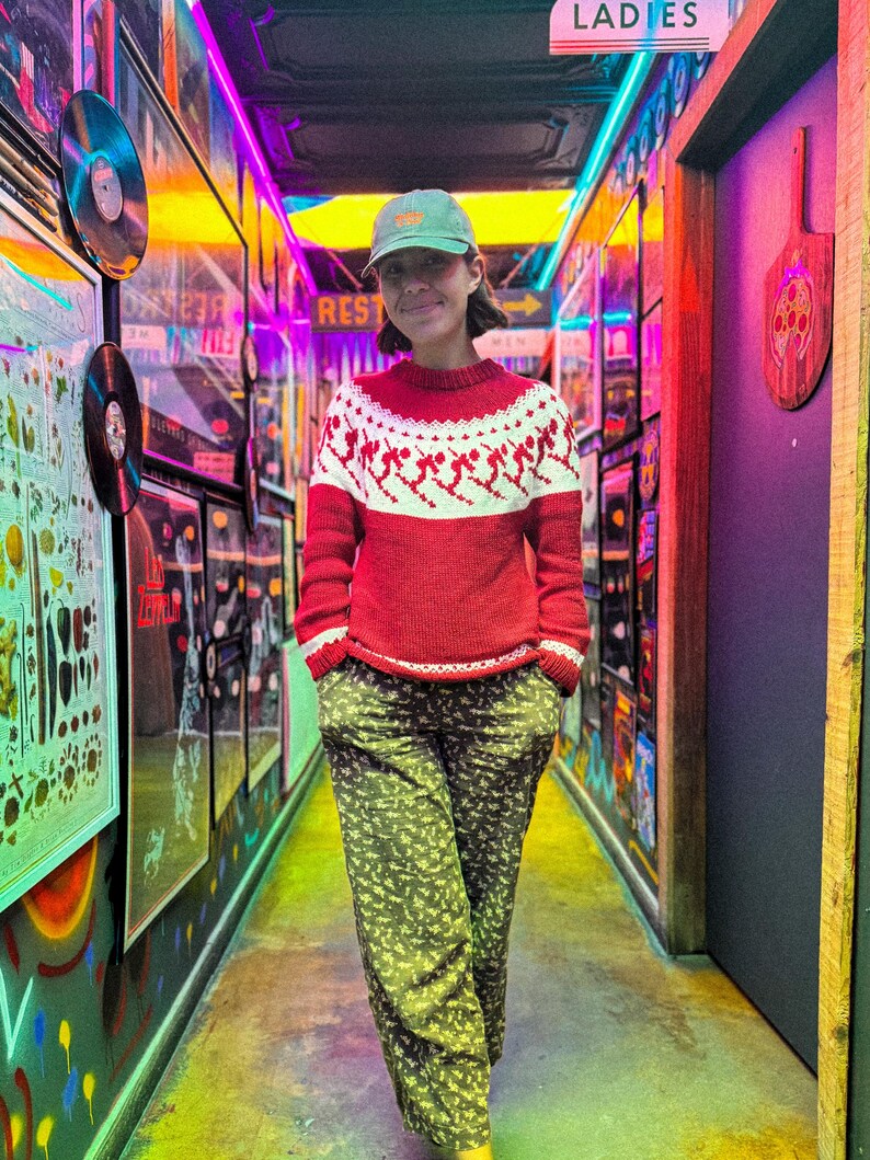 Puede incluir: Una persona con un su&eacute;ter rojo y blanco con tem&aacute;tica de esqu&iacute;, pantalones verdes con estampado floral y una gorra verde. La persona est&aacute; en un pasillo con luces de ne&oacute;n coloridas y obras de arte enmarcadas. Un letrero arriba dice "Ladies".