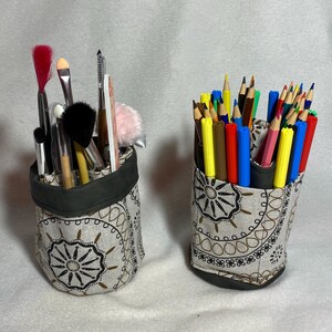 Peut inclure: Deux porte-crayons en tissu avec un motif décoratif. L'un contient des pinceaux de maquillage, l'autre des crayons de couleur. Les supports ont une bordure et une base vert foncé, et le tissu a un fond beige avec un motif marron et noir.