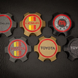 May include: A collection of automotive wheel center caps featuring the Lexus and Toyota logos. The caps are in various colors, including black, gold, and gray, with red and orange accents. Some caps have Japanese text.
