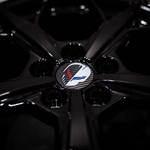 以下が含まれることがあります： 光沢のある黒い車のホイールのクローズアップ。ホイールのセンターキャップには、赤、白、青のアクセントが付いたロゴがあります。ホイールのデザインには、複数のスポークと中央のハブが含まれています。