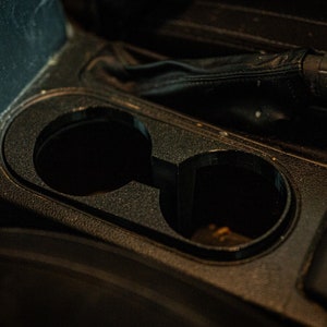 以下が含まれることがあります： 黒い車のセンターコンソールをクローズアップした写真。カップホルダーが2つあり、空になっています。コンソールはテクスチャのある素材でできています。