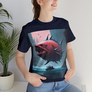 May include: A navy blue t-shirt with a graphic print of a large, red, toothy fish with sharp fins flying over a body of water. The fish is being held by a rope attached to a small boat.