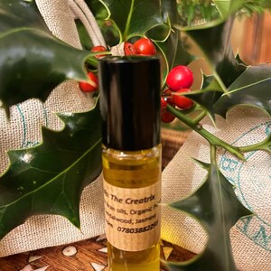 May include: A small glass bottle with a black cap and a yellow liquid inside. The bottle is labeled "The Creatrix" and lists "Essential oils, Organic Patchouli, Sandalwood, Jasmine" and a phone number. The bottle is sitting on a wooden surface with green leaves and red berries in the background.