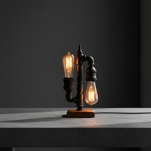 Könnte beinhalten: Industriell gestaltete Tischlampe mit schwarzem Rohrrahmen und zwei Edison-Glühbirnen. Die Lampe steht auf einem rechteckigen Holzsockel. Die Glühbirnen strahlen ein warmes Licht aus, ideal für eine Vintage-Note. Höhe ca. 30 cm.