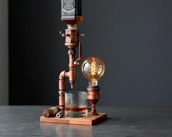 May include: A copper pipe lamp featuring an inverted Jack Daniel's bottle as a dispenser, a glass tumbler filled with amber liquid, and an Edison bulb. The assembly is mounted on a rectangular wooden base with a cigar and a metal tube accessory.