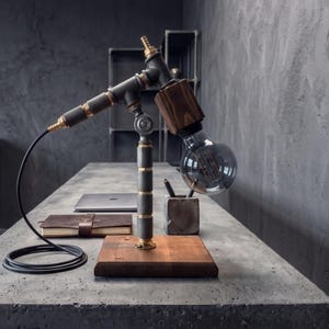 May include: Industrial-style desk lamp with a wooden base and a unique design. The lamp features a large, clear light bulb and metal piping with brass accents. A black cord extends from the lamp, adding to its industrial aesthetic. The lamp sits on a concrete desk.