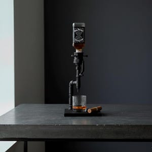 May include: A black metal liquor dispenser with a glass of amber liquid and a cigar on a dark surface. The dispenser holds a bottle of Jack Daniel's whiskey. The setup is on a gray table against a dark background.