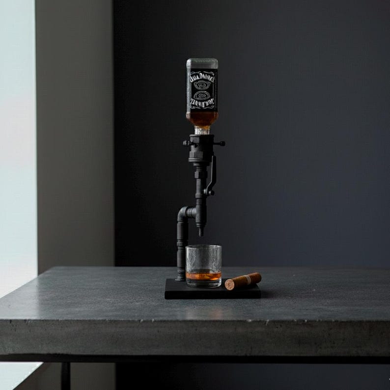 May include: A black metal liquor dispenser with a glass of amber liquid and a cigar on a dark surface. The dispenser holds a bottle of Jack Daniel's whiskey. The setup is on a gray table against a dark background.