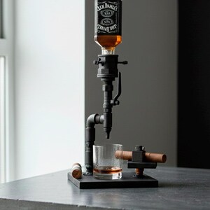 May include: A black metal liquor dispenser with a Jack Daniel's bottle, a glass of amber liquid, and a cigar holder. The dispenser has a pipe-like design and sits on a black base. A cigar rests in the holder.
