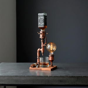 May include: A copper-toned liquor dispenser with a vintage Edison bulb and a glass of whiskey. The dispenser holds a bottle of Jack Daniel's whiskey and sits on a wooden base. A cigar rests on the base.