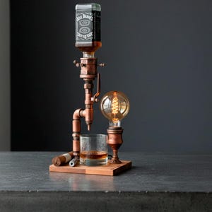 May include: A copper-toned whiskey dispenser with a vintage industrial design. It features a Jack Daniel's bottle, a glass of whiskey, a light bulb, and a wooden base. A cigar and a cigar cutter are also present.