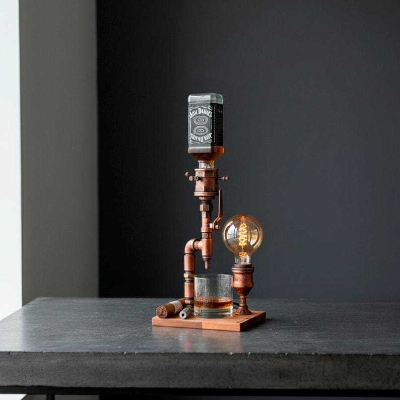 May include: A copper-colored liquor dispenser with a glass and a light bulb. The dispenser holds a bottle with a black and white label. A glass of amber liquid sits below the dispenser. A cigar rests on the wooden base.