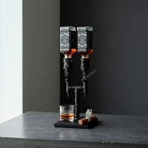 May include: A black metal liquor dispenser with two bottles of whiskey. A glass of whiskey, a cigar, and a lighter sit on a black base. The dispenser is on a gray surface against a dark gray background.
