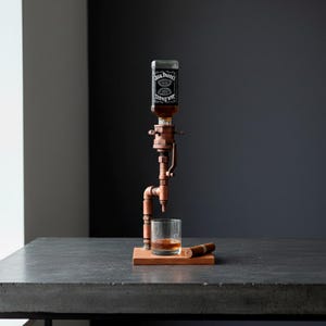 May include: A copper-toned liquor dispenser with a Jack Daniel's bottle, a glass of amber liquid, and a cigar on a wooden base. The dispenser has a pipe-like design and sits on a dark gray surface.