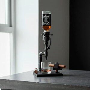 May include: A black metal liquor dispenser with a Jack Daniel's bottle. The dispenser has a pipe-like design with a glass of amber liquid below the spout. A copper-colored piece sits on a small black base.
