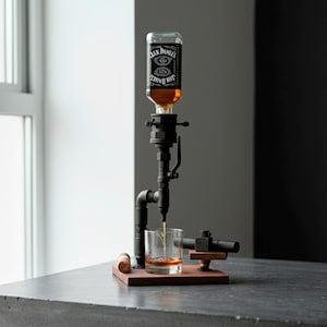 May include: A black metal liquor dispenser with a Jack Daniel's bottle attached. The dispenser is pouring amber liquid into a glass. The setup includes a wooden base and a small wooden stopper.