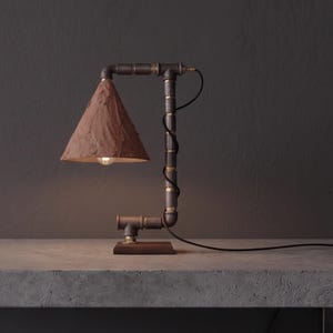 May include: Industrial-style table lamp with a brown paper shade. The lamp features a metal pipe frame with brass fittings and a wooden base. A black cord extends from the lamp. The shade is cone-shaped and directs light downward.