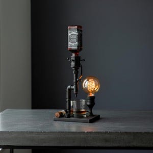 May include: A unique bar accessory featuring a Jack Daniel's bottle dispenser, a vintage-style light bulb, and a rocks glass. The dispenser is made of black metal pipes and sits on a dark wooden base. A cigar rests on the base.