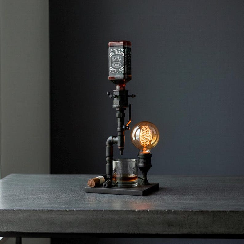 May include: A unique bar accessory featuring a Jack Daniel's bottle dispenser, a vintage-style light bulb, and a rocks glass. The dispenser is made of black metal pipes and sits on a dark wooden base. A cigar rests on the base.