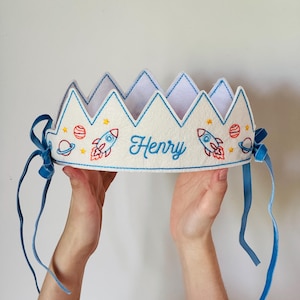May include: A white felt crown featuring embroidered space-themed designs including rockets, planets, and stars in red, blue, and yellow. The name Henry is embroidered in blue script in the center. The crown has blue ribbon ties on both sides.