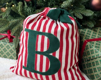 May include: A festive Christmas gift bag with red and white stripes, a green bow, and a large green letter "B". The bag is placed on a white surface, with a Christmas tree in the background.