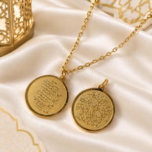 May include: A gold-toned necklace featuring a circular pendant with engraved Arabic calligraphy, accompanied by a matching circular charm also displaying intricate Arabic script. The jewellery pieces have a polished finish and are connected by a delicate chain.