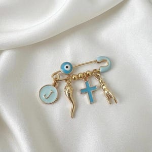 May include: Gold-tone safety pin with multiple charms. Charms include a blue evil eye, a gold chili pepper, a blue cross, a gold hand, and a light blue circle with the letter "J". The pin and charms are set against a white background.