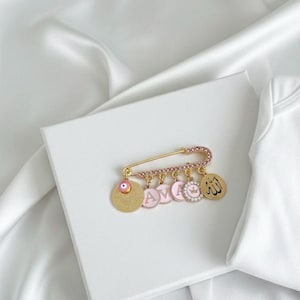 May include: A gold-tone safety pin with pink detailing and charms. The pin has pink crystal embellishments and hanging charms, including a coin with an eye, pink circles with the letters "AVA", a crown, and a charm with Arabic script.