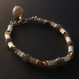 Könnte beinhalten: Ein silbernes und goldenes Armband mit einem Perlenanhänger und grauen Edelsteinperlen.