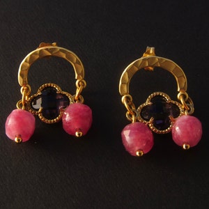 May include: Gold hoop earrings with a dangling clover charm featuring a purple gemstone and two pink gemstone beads.