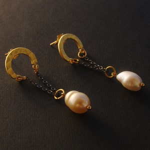 Peut inclure: Une paire de boucles d'oreilles en or avec un design de cerceau martelé et une perle pendante. Les cerceaux sont reliés à une chaîne noire qui se termine par une seule perle.