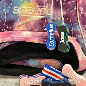 Könnte beinhalten: Nahaufnahme von sechs bunten Pool-Schwimmnudeln mit darauf geschriebenen Namen. Die Schwimmnudeln sind rosa, blau, schwarz, grün und rot. Die Namen auf den Schwimmnudeln sind Cornelius, Jose, Reagan, Johnny und Mollie.