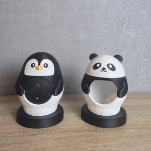 Könnte beinhalten: Zwei dekorative Tierhalter. Einer ist ein Pinguin mit schwarzem Körper, weißem Bauch und orangefarbenem Schnabel. Der andere ist ein Panda mit weißem Kopf, schwarzen Ohren und schwarzen Akzenten. Beide stehen auf schwarzen Sockeln.