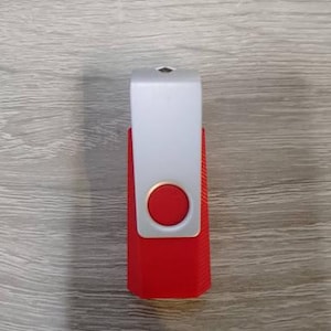 May include: A red and white USB flash drive. The drive has a rectangular red base with a silver top. A red circle is centered on the silver portion. The drive is on a wooden surface.