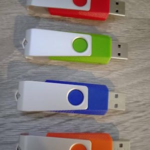 Puede incluir: Cuatro memorias USB en rojo, verde, azul y naranja. Cada unidad tiene una cubierta blanca con un bot&oacute;n de color y un conector USB. Las unidades est&aacute;n dispuestas verticalmente sobre una superficie de vetas de madera clara.