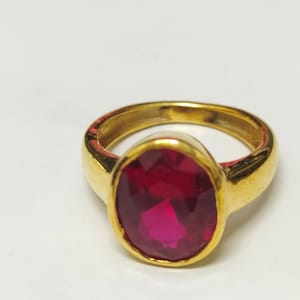 Puede incluir: Un anillo de oro con una gran gema roja oscura de corte ovalado. El anillo tiene una banda de oro gruesa y pulida y un engaste sencillo. La gema es el punto focal, captando la luz y mostrando un color vibrante.
