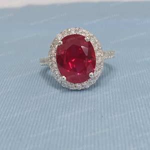 May include: A silver ring with a large oval-shaped ruby gemstone surrounded by a halo of smaller diamonds. The ring is set on a blue background.