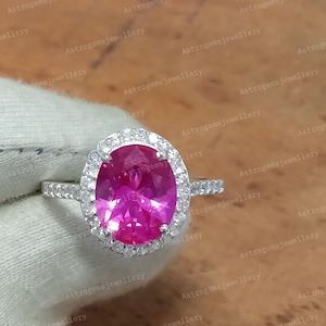 Könnte beinhalten: Ein silberner Ring mit einem großen ovalen rosafarbenen Edelstein, der von einem Heiligenschein aus kleineren weißen Edelsteinen umgeben ist.