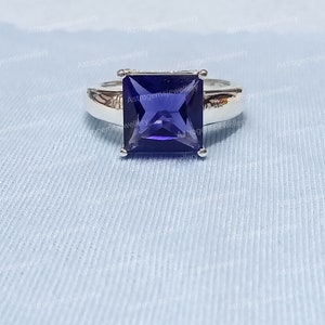 May include: A silver ring featuring a square-shaped, deep purple gemstone. The ring is displayed against a white fabric backdrop.