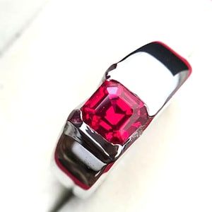 May include: A silver ring featuring a vibrant, square-cut, ruby-red gemstone. The ring's band has a polished, reflective surface, creating a striking contrast with the deep red of the stone. The design is simple yet elegant.