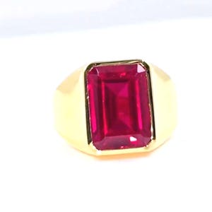 May include: A gold ring with a large, rectangular, deep red gemstone. The ring has a simple, classic design with a polished gold band and a bezel setting. The gemstone is a vibrant, saturated red. The ring is a statement piece.