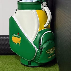 May include: A green, white, and yellow golf bag filled with white golf balls. The bag features the Masters logo, a map of the United States, and the text A TRADITION UNLIKE ANY OTHER SINCE 1934 embroidered on the front pocket with black zippers.