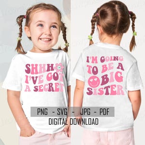 Op de afbeelding: Een wit T-shirt met roze en rode tekst die "SHHH I'VE GOT A SECRET" op de voorkant en "I'M GOING TO BE A BIG SISTER" op de achterkant zegt. Het shirt wordt gedragen door een kind.